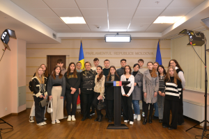 Eine Gruppe von Menschen steht vor einem Podium mit Mikrofon, umgeben von Fahnen, einer Wand mit dem Text "Moldova Youth Congress 2018", einer Tür, einem Dach mit Deckenleuchten und Ständern mit Leuchten.