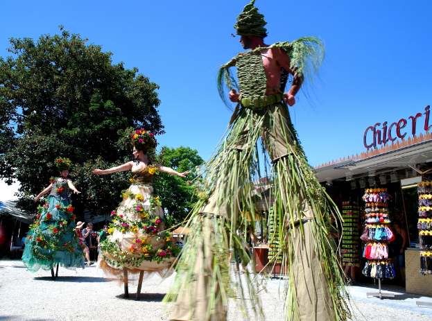 Drei Personen in Kostümen tanzen in der Mitte, mit Geschäften, die Kleidung und Gegenstände im Hintergrund anbieten, Text auf einem Gebäude, zusätzliche Geschäfte und Bäume links, ein Gehweg unten und Himmel oben.