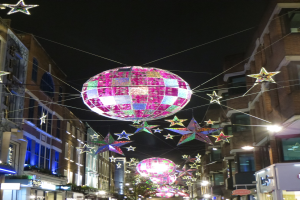 Eine festlich geschmückte Stadtstraße bei Nacht mit zahlreichen Lichtern und Dekorationen, darunter ein großer Diskokugel an einem Gebäude.