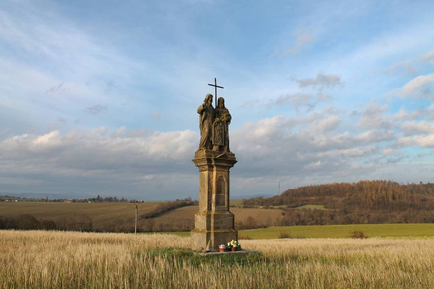 Ein Denkmal mit einem Kreuz darauf steht in einem Feld umgeben von Pflanzen und Blumen, mit Bäumen und Pfählen im Hintergrund, als Mahnmal für Holocaust-Opfer in Deutschland.