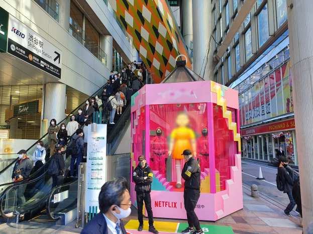 Eine Gruppe von Menschen steht vor einem Gebäude mit Glaswänden und Texttafeln, einige tragen Masken, während eine Rolltreppe mit Menschen daneben fährt; ein pinker Kasten in der Mitte zeigt Schaufensterpuppen und trägt den Text "Netflix's neuer Netflix-Stand in Tokyo."