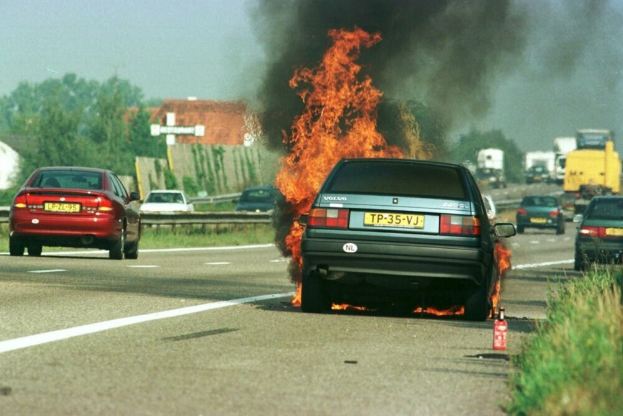 Ein Auto in Flammen auf dem Seitenstreifen mit umliegenden Fahrzeugen, B%C3%A4umen, Geb%C3%A4uden und einem klaren blauen Himmel im Hintergrund und einem Feuerl%C3%B6scher in der N%C3%A4he auf dem Gras.