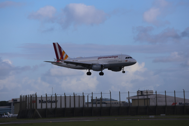 Germanwings Airbus A320-200 beim Start vom Frankfurt Airport, mit Gras darunter, einem Zaun im Vordergrund, Gebäuden und Bäumen im Hintergrund und einer bewölkten Himmel