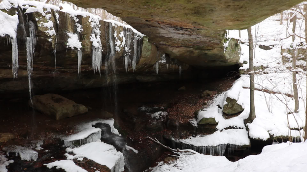 Ein kleiner Wasserfall ergießt sich über einen schneebedeckten, steinigen Hang in einem bewaldeten Gebiet, mit Eiszapfen an den Felsen.