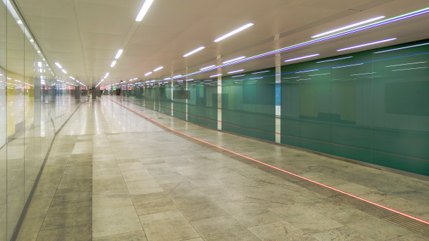 Ein langer Gang mit Glaswänden auf beiden Seiten und einer beleuchteten Decke, mit ein paar Menschen, die hindurchgehen und so einen belebten Eindruck einer Stadt erwecken.