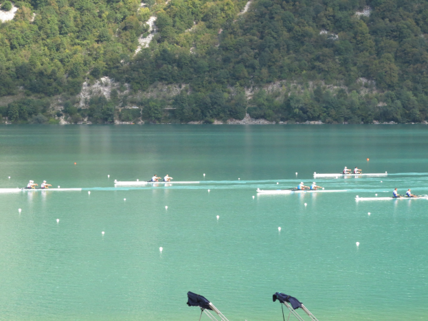 Eine Gruppe von Menschen, die bei einem Wettbewerb auf einem Gewässer mit Bäumen und einem Hügel im Hintergrund rudern, mit Kleidung, die über Stände am Boden gehängt ist.