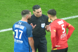 Schiedsrichter spricht mit zwei Männern auf einem Fußballfeld, einer trägt ein schwarzes Hemd und Hose, der andere ein blaues und rotes T-Shirt mit Text, beide tragen eine Brille.