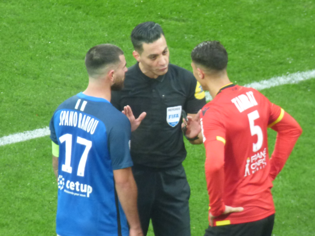 Schiedsrichter spricht mit zwei Männern auf einem Fußballfeld, einer trägt ein schwarzes Hemd und Hose, der andere ein blaues und rotes T-Shirt mit Text, beide tragen eine Brille.