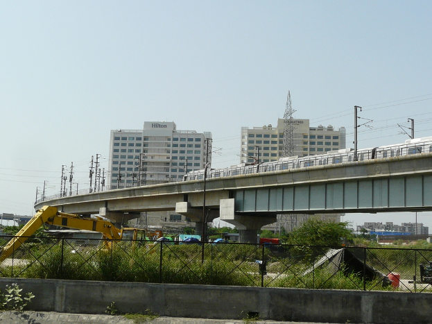 Ein klarer Himmel mit Gebäuden unten, eine Brücke mit einem darüberfahrenden U-Bahn-Zug, Strommasten, die durch Drähte verbunden sind, ein Pflugfahrzeug darunter, Pflanzen und Bäume, ein schwarzes Zelt und ein Zaun.