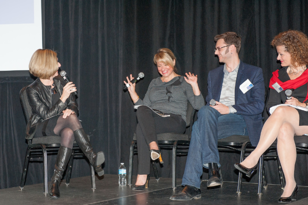 Ein Mann und drei Frauen sitzen auf Stühlen, halten jeweils ein Mikrofon und lächeln, mit einer Flasche und einem Bildschirm im Hintergrund und Vorhängen dahinter.