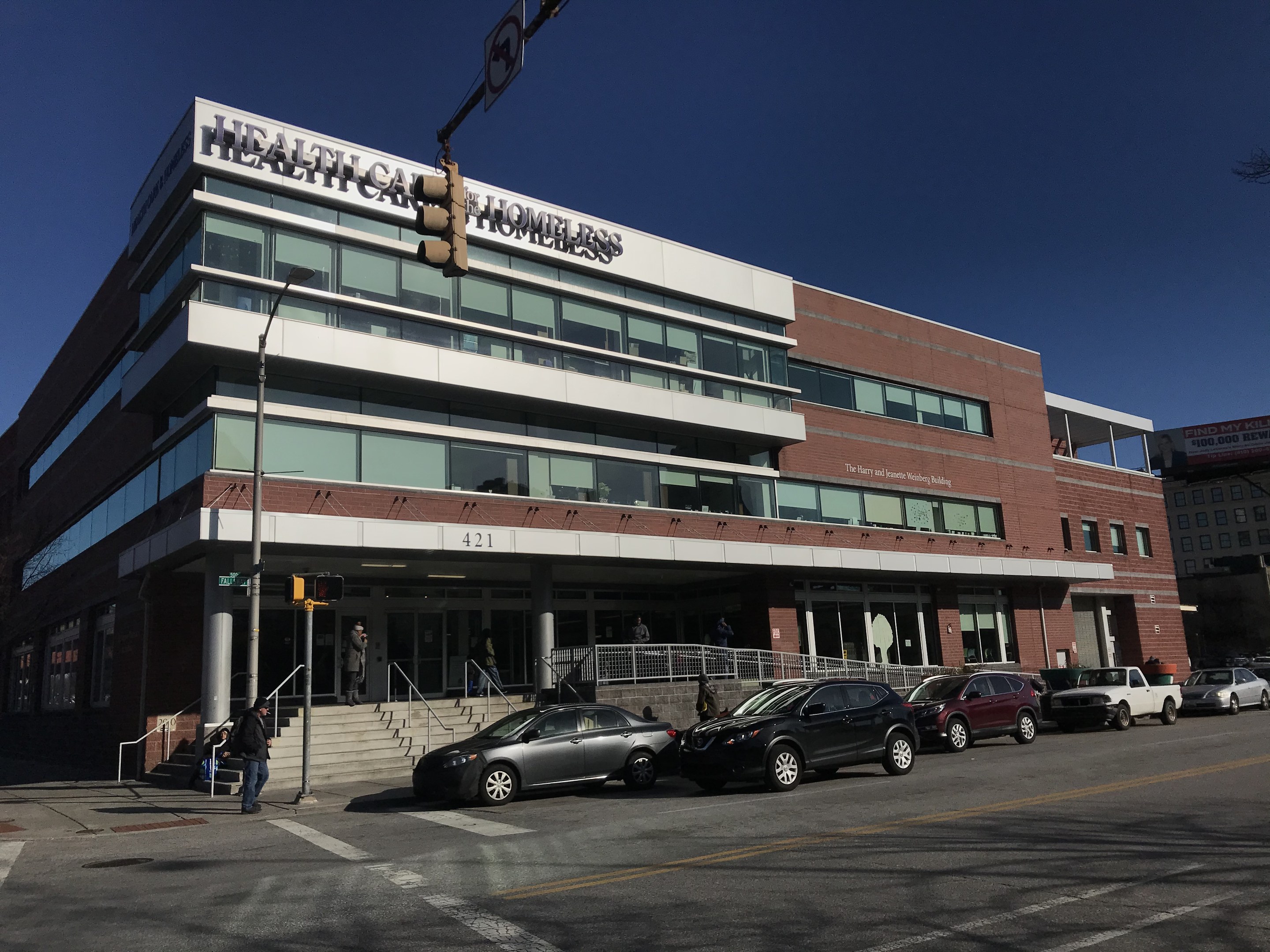 Großes Gebäude mit der Aufschrift "New York City Health Care Homeless Center" mit geparkten Autos, Fußgängern, Straßenmöbeln und Bäumen unter einem klaren Himmel.