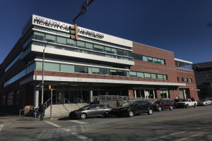 Großes Gebäude mit der Aufschrift "New York City Health Care Homeless Center" mit geparkten Autos, Fußgängern, Straßenmöbeln und Bäumen unter einem klaren Himmel.