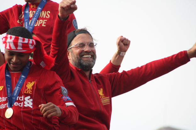 Liverpool-Trainer Jürgen Klopp feiert mit seinen Spielern den Sieg in der UEFA Champions League, während er eine rote Jacke trägt und von zwei medaillen tragenden Personen umgeben ist. Der Himmel ist im Hintergrund zu sehen.