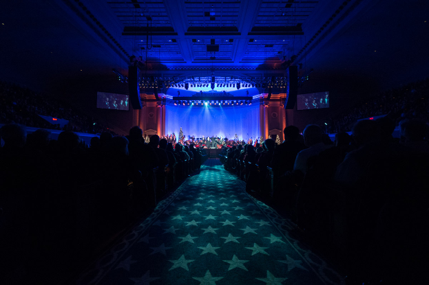 Ein großer Auditorium mit sitzenden Menschen, eine Bühne mit Musikern, die Instrumente spielen, und eine Decke mit Lichtern und Fernsehgeräten, was einen Konzertsaal für die Nationale Convention 2019 vermuten lässt.