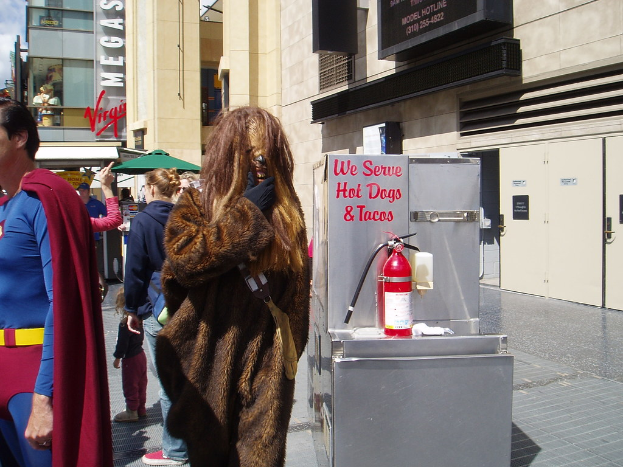 Eine Menschenmenge, darunter einige in Kostümen, versammelte sich vor einem Gebäude mit sichtbarer Beschriftung und Türen, mit einer Box und einem Feuerlöscher in der Nähe.