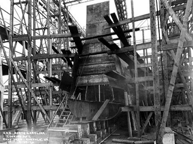 Schwarzes und weißes Foto eines Schiffs in einem Schiffbauwerk, mit hölzernen Pfählen und Treppen im Vordergrund und Himmel im Hintergrund, mit Text "Marinebewaffnung" in der linken unteren Ecke.