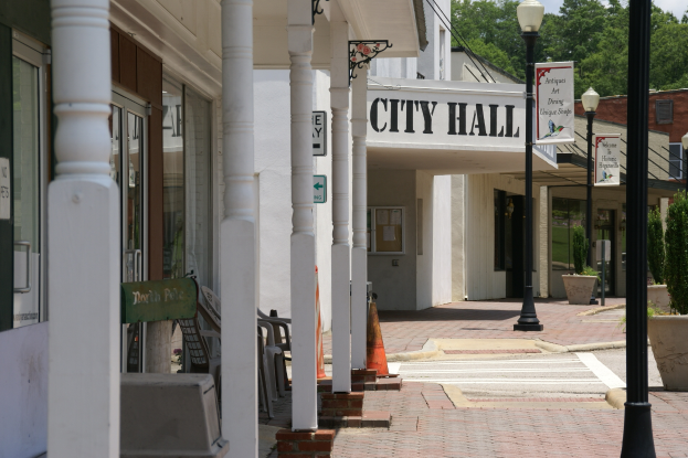 Ein Rathaus umgeben von Gebäuden, Straßenmasten, Straßenlaternen, Namensschildern, Kraftfahrzeugen, Absperrgittern, Stühlen, Zimmerpflanzen, Bäumen und einem bewölkten Himmel.