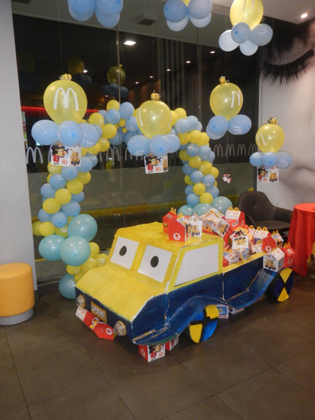 Eine McDonald's-Geburtstagsfeier mit einem gelben und blauen Lastwagen, der mit Ballons und anderen Dekorationen geschmückt ist, einem roten Tisch mit Gegenständen, Hockern und einer Glaswand im Hintergrund unter festlicher Deckenbeleuchtung.
