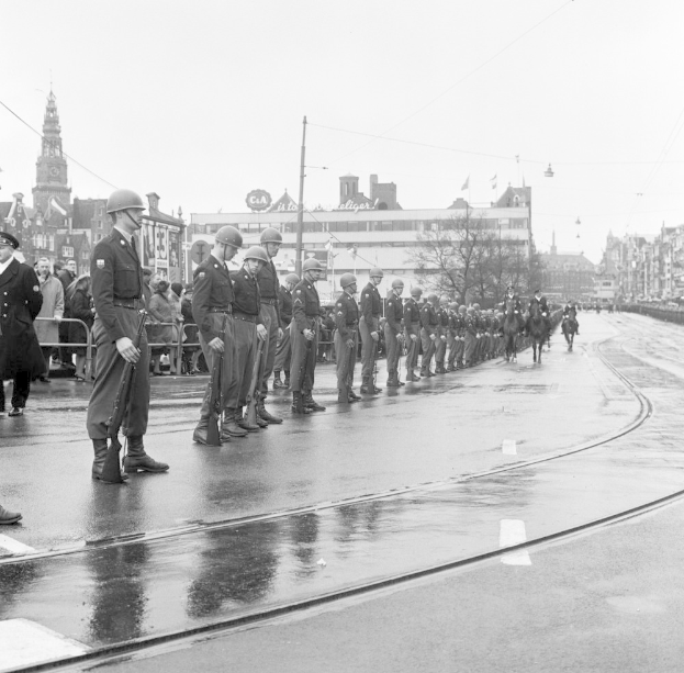 Eine Gruppe von Polizisten in Helmen und mit Waffen in der Hand steht an der Straße, hinter ihnen ein Zaun und im Hintergrund Gebäude, Bäume, Pfosten, Drähte, Flaggen und der Himmel, dargestellt in Schwarz-Weiß.