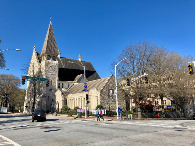 St. Luke's Episcopal Church, eine große Kirche mit einem Turm, steht an einer Straßenecke umgeben von Gebäuden, Fahrzeugen, Fußgängern und Bäumen unter einem klaren blauen Himmel.