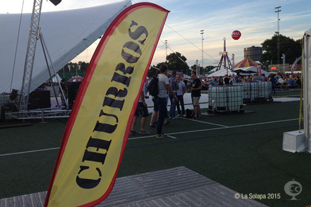 Eine bunte KarnevalsSzene mit Zelten, einem zentralen Mast, Menschen, Bäumen, Strommasten, Straßenbahnen auf der Straße und einem Eisenständer auf der linken Seite unter einem klaren Himmel.