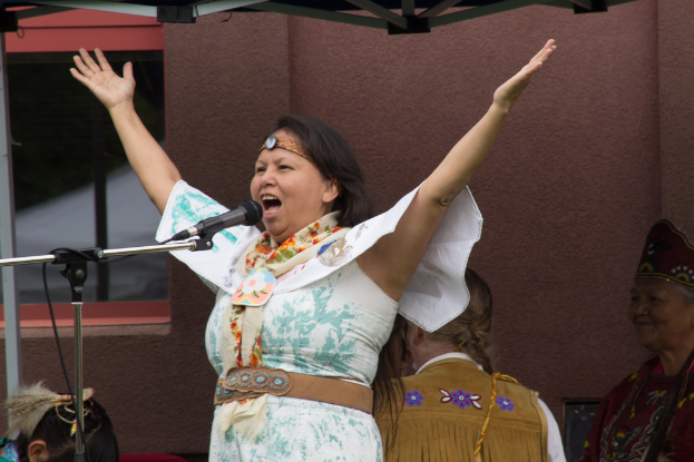 Eine Frau in einem weißen Kleid steht auf einer Bühne und schreit in ein Mikrofon, mit einer Wand und einem Eisenrahmen im Hintergrund.