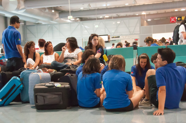 Ein belebter Flughafeninnenraum mit vielen Menschen, die auf Flüge warten, Gepäck auf der linken Seite und Glastüren mit Richtungsschildern im Hintergrund.