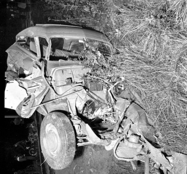 Ein Schwarz-Weiß- Foto eines schwer beschädigten Autos am Straßenrand, mit einer zerquetschten Front, zersplitterten Fenstern, einem Baum im Hintergrund und verstreuten Gegenständen um die Unfallstelle.
