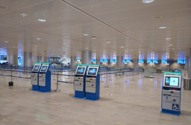 Ein geräumiger Flughafen-Check-in-Bereich mit zahlreichen blauen und weißen Maschinen, Säulen mit Bändern, Deckenleuchten und verschiedenen anderen Gegenständen.