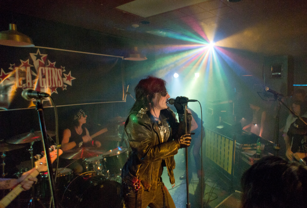 Eine Frau in einer schwarzen Jacke singt in ein Mikrofon auf der Bühne, während ein Mann in Sonnenbrille und schwarzem Stirnband hinter ihr die Bassgitarre spielt, unter Diskoleuchten und einem 'LA Guns'-Plakat.
