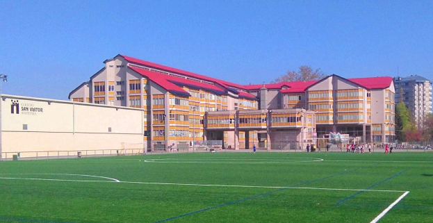 Ein Fußballfeld mit einem großen Gebäude im Hintergrund, das als San Visitor Academy beschriftet ist, umgeben von Bäumen und Laternen, ein paar Menschen auf dem Feld und der Himmel darüber sichtbar.