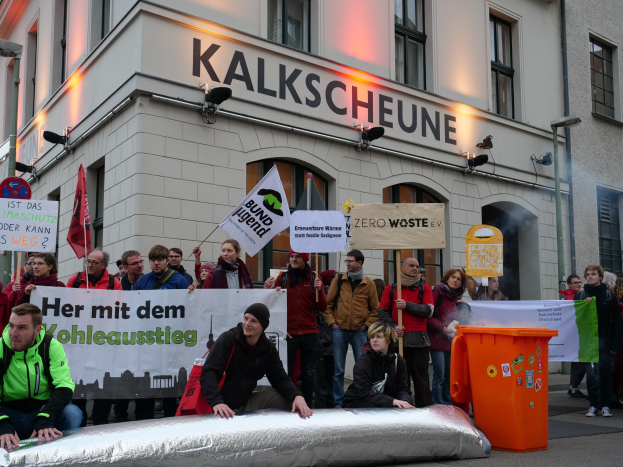 Eine Gruppe von Menschen mit Transparenten und Schildern steht vor einem Gebäude, mit zwei Personen im Vordergrund und einem Müllcontainer auf der rechten Seite, während einer Protestaktion in Deutschland, mit Gebäuden, Fenstern, Lichtern und Schildern im Hintergrund.
