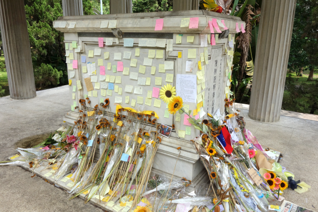 Ein Gedenkstein in einem Park, geschmückt mit Post-it-Zetteln und Blumen, umgeben von Bäumen, Pflanzen und Gras.