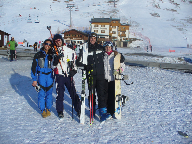 Vier Personen posieren mit Skate-Pads, Werkzeugen und Gebäuden im Hintergrund vor einer schneebedeckten Berglandschaft.