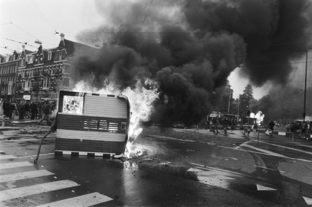 Ein Schwarz-Weiß-Foto eines brennenden Busses am Straßenrand mit Menschen, Pfählen, einem Schild, Gebäuden und einem bewölkten Himmel im Hintergrund.