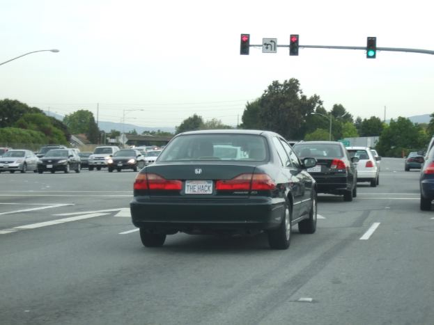 Eine befahrene Straße mit mehreren Fahrzeugen, drei Strommasten und einem Verkehrszeichen auf der rechten Seite unter einem klaren Himmel.