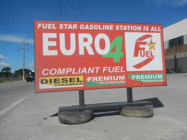 Eine Tankstelle mit einem "Euro4 Fuel"-Schild im Vordergrund, Fahrzeuge auf der Straße, Strommästele mit Drähten, Bäume, ein Gebäude auf der rechten Seite und ein sichtbarer Himmel.