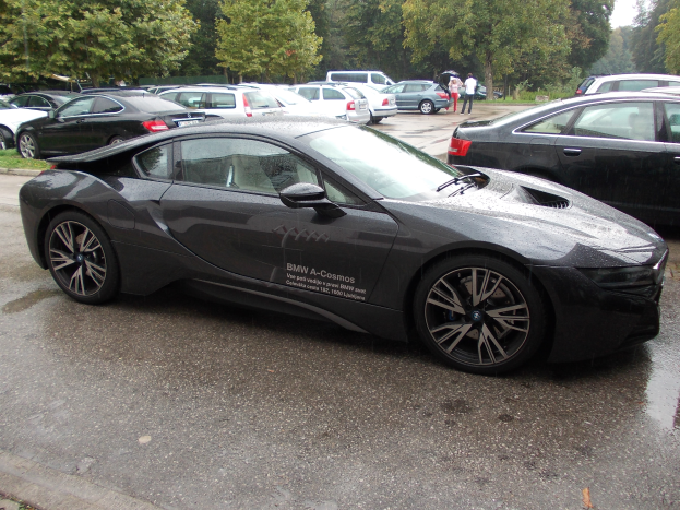 Ein schwarzer BMW i8 auf einem Parkplatz neben anderen Autos geparkt, Menschen in der Nähe, Bäume und einen klaren blauen Himmel im Hintergrund.