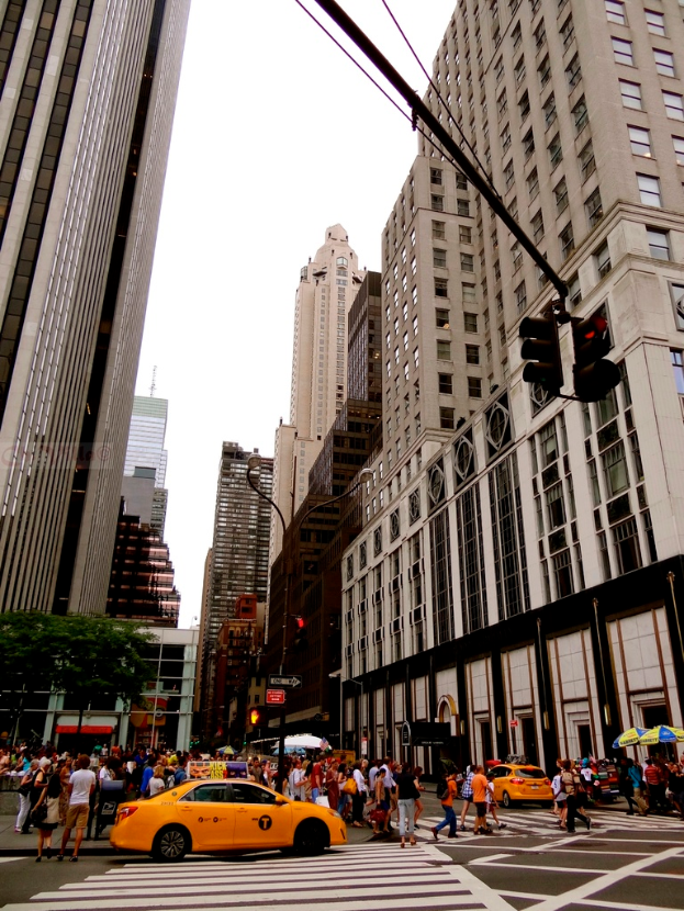 Eine Stadtansicht mit Gebäuden, Ampeln, Fenstern, einigen Fußgängern, Autos und dem Himmel.
