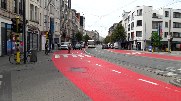 Eine Stadtstraße mit einem roten Fahrradweg, Fahrzeuge auf der Straße, Fußgänger auf dem Gehweg, Gebäude mit Fenstern auf beiden Seiten und Bäume mit einem klaren blauen Himmel im Hintergrund.