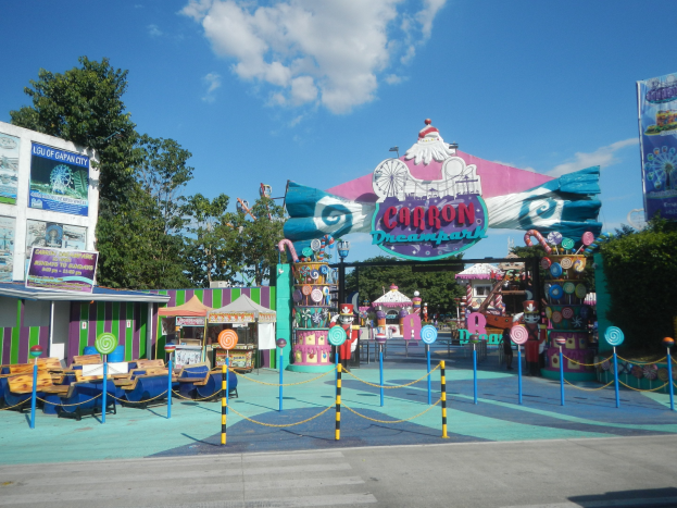 Karussell am Eingang eines Parks, mit Straße, Umzäunung, Buden, Banner, Bäumen und bewölktem Himmel im Hintergrund.