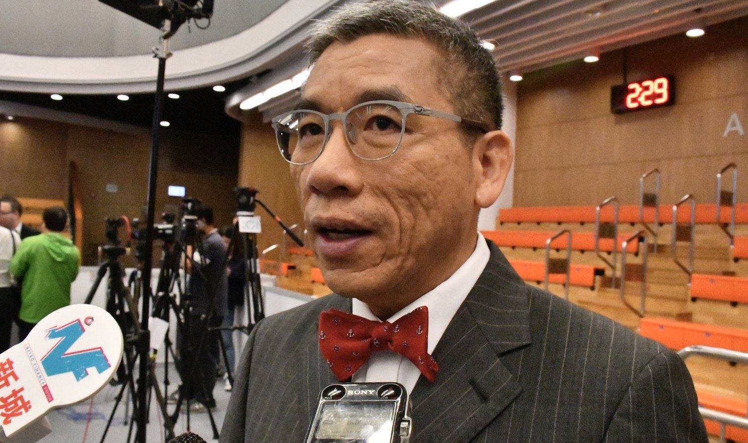 Ein Mann im Anzug und mit Schleife spricht zu Reportern und hält ein Mikrofon, umgeben von Kameras, Stativen, Bänken, einer Digitaluhr und Scheinwerfern im Hintergrund.