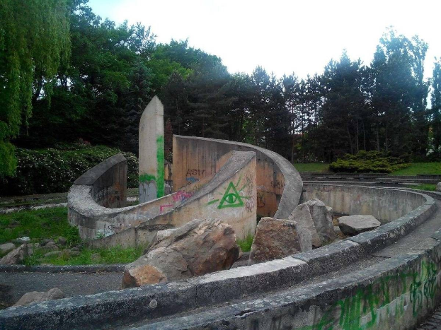 Verlassener Skatepark mit Graffiti an seiner Seite, umgeben von Felsen, Gras, Pflanzen und Bäumen unter einem klaren blauen Himmel.