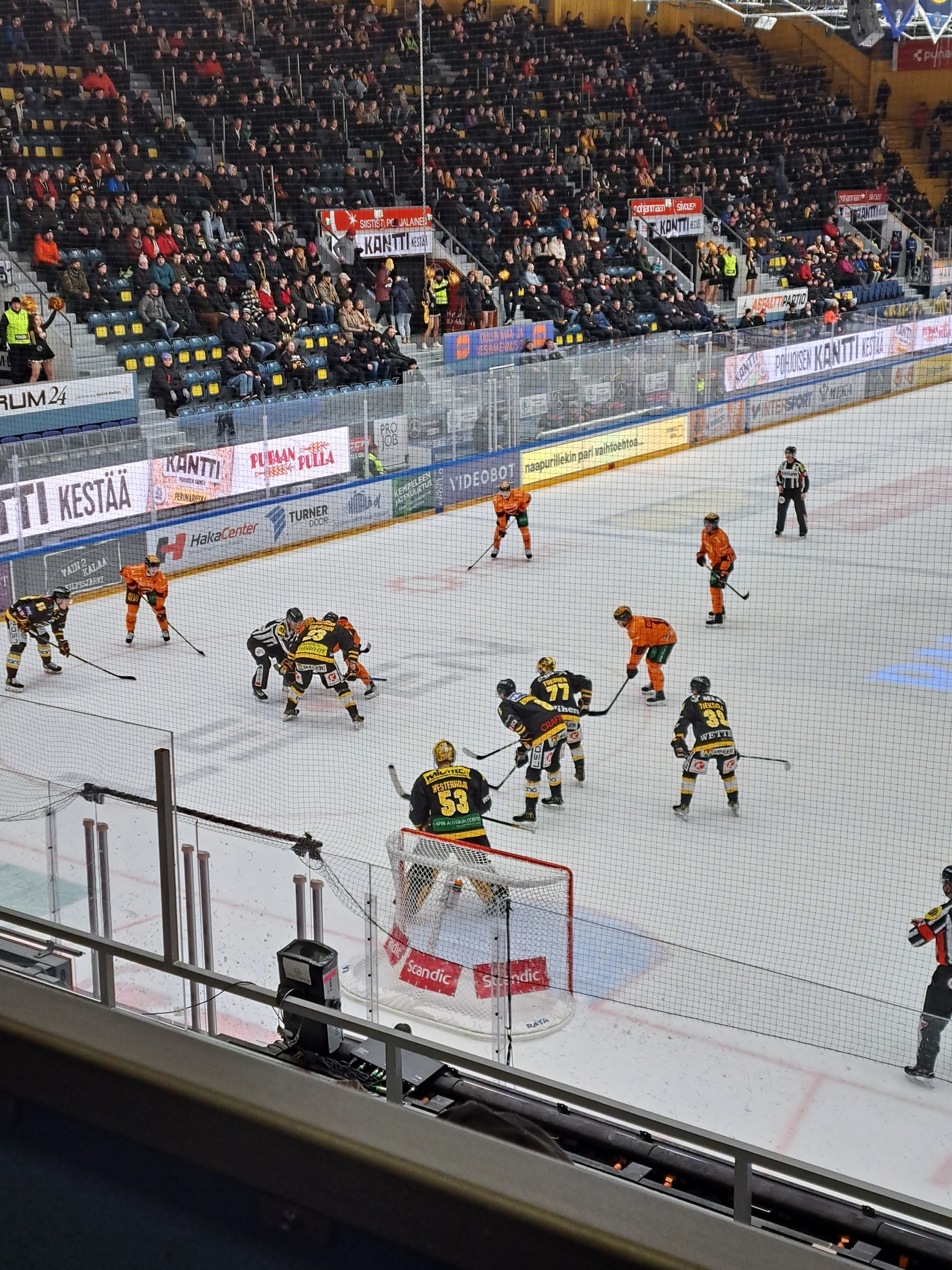 Eine Gruppe von Menschen, die Hockey auf einem Eisstadion spielen, mit einem Netz, einer Umzäunung, sitzenden Zuschauern, Bannern und Scheinwerfern von oben.