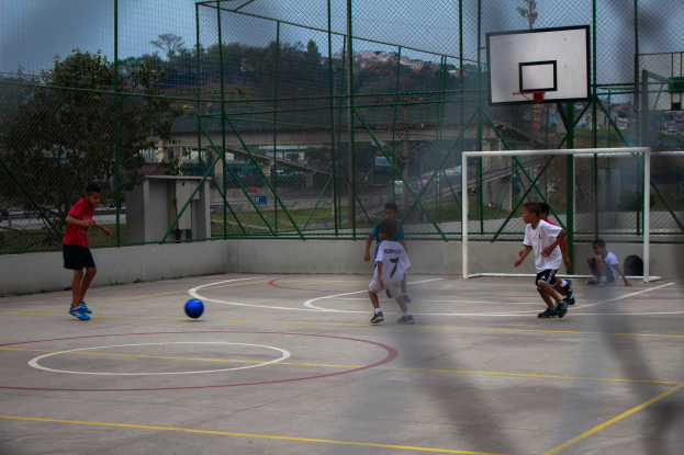 Junge Jungs spielen Basketball auf einem Outdoor-Court mit einem Zaun, einem Ball im Spiel, Bäumen, Gebäuden und einem klaren blauen Himmel im Hintergrund.