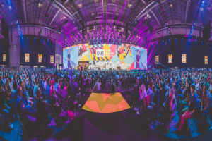 Eine große Menschenmenge sitzt auf Stühlen vor einer hell erleuchteten Bühne mit mehreren stehenden Personen, wahrscheinlich bei einem Online-Marketing-Rockstars-Event, umgeben von Bildschirmen.