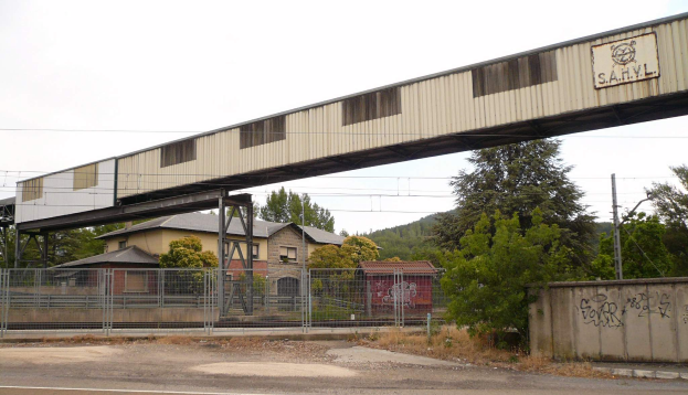 Eine Eisenbahnbrücke spannt sich über eine Straße mit Graffiti, umgeben von einem Metallzaun, mit Bäumen, Häusern, Strommasten und einem bewölkten Himmel im Hintergrund.
