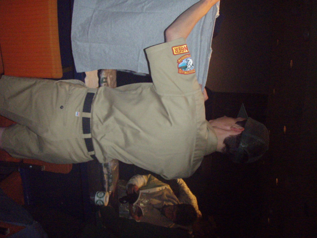 Ein Polizeibeamter hält ein T-Shirt in den Vordergrund, mit einem Jungen im Hintergrund, der ein Buch und einen Becher hält, der neben einem Schreibtisch und Lampen steht.