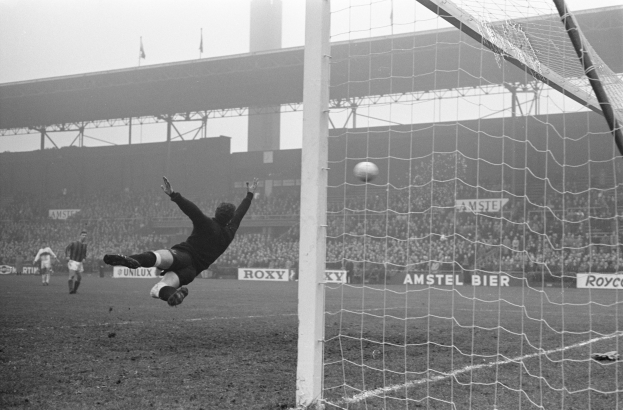 Ein Schwarz-Weiß-Foto eines Fußballspielers, der vor einem Tor einen Ball hält, mit einem Netz auf der rechten Seite und einer Menge Menschen im Hintergrund, sowie Schildern mit Text, einer Brücke und Fahnen mit Stangen.