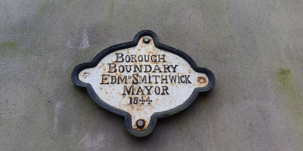 Metallplakette an einer Gebäudeseite mit schwarzem Hintergrund und weißer fettgedruckter Schrift, die "Borough Boundary Edm Smithwick Mayor" liest.